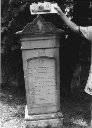 Grabstein 169 (Index: 100110651) Material: Sandstein Zustand: Giebel beschädigt, ehemals schwarz ausgelegt Schrift Maße HxBxT: 167x64x36 cm Beschreibung: Stele, Sockel profiliert, Giebel mit mehrfach geschwungenem Abschluß, Schriftfeld eingetieft mit Segmentbogenabschluß Symbolik: florale Motive: Grabstein 169