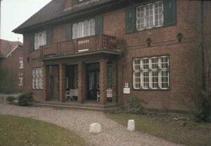 Kirchstraße 10: traufständiges Backsteinhaus mit Walmdach: Straßenfront mit Pfeilerportikus, Eingangstür, Altan, Gaube mit Bogendach: davor Auffahrt