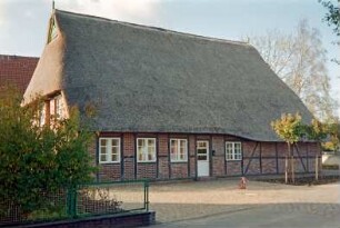 Reinbeker Straße 4a: giebelständige Fachwerkkate mit Reetdach: Traufseite, von links Wohntrakt, Eingangstür, Sprung, Wirtschaftstrakt: links Giebelseite Wohntrakt [halb verdeckt], oben Giebelzier, Giebelpfahl: davor Hof mit Pflasterung, junge Bäume, Grundstückszaun: im Vordergrund Stichstraße