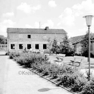 Ahrensburg: Reeshoop: Außenaufnahme: links Wirtschaftsgebäude, rechts Wohnflügel: Grünanlage mit Parkbänken, Parklampe, Bäumen, Sträuchern: vorne links Zufahrt