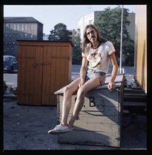 Junger Mann in Shorts, auf einer Kiste sitzend, T-Shirt „Just passin’ thru“ von Robert Crump