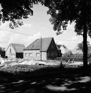 Volksschule: dahinter Häuser: im Vordergrund links Fundamente und Grundmauern eines Neubaus: rechts Baum