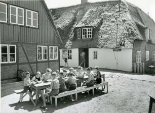 Hallig Süderoog. Schulausflug. Eine Jungengruppe beim Essen im Freien. Im Hintergrund reetgedeckte Häuser des Bauernhofes