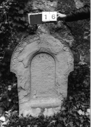 Grabstein 16 (Index: 99793030) Material: Sandstein Zustand: sehr stak verwittert, Texttafel fehlt Maße HxBxT: 113x70x19 cm Beschreibung: Stele, überstehender Rundbogenabschluß, Schriftfeld eingetieft mit Rundbogenabschluß, eingesetzte Texttafel Symbolik: Band aus floralen Motiven: Grabstein 16