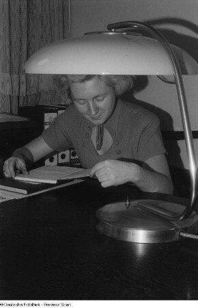 Blick auf eine am Schreibtisch sitzende Frau; im Vordergrund eine Schreibtischlampe