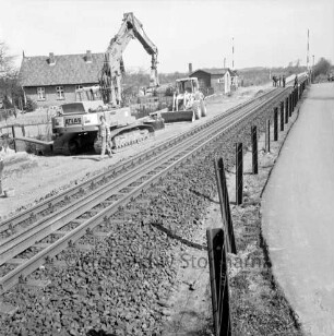 Bahnübergang bei Schrankenposten 60, Im Seybeck: Bahnstrecke zwischen Hamburg und Lübeck: Gleiserneuerung: Raupenbagger, Schaufellader, Schienenstücke, Arbeiter: hinten Schrankenwärterhaus, Schranke geöffnet, Personen auf Übergang: hinten links Wohnhaus, Garten: rechts Zaun, Straße
