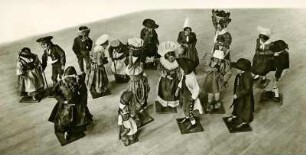 Siebzehn handgefertigte Spielfiguren in traditioneller Stoffkleidung im Gruppenbild auf einer Holzplatte. Theodor-Zink-Museum. Totale