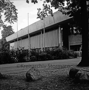 Stormarnhalle: Am Bürgerpark: vorn Findling, Baum und Weg: dahinter Zaun, Büsche und drei Fahnenmasten
