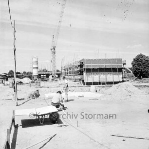 Langnese: Gewerbegebiet: Hammoorer Weg: Honigfabrikation: Betriebsgebäude in Bau: Rohbau mit Gerüsten, Baumaterialien: Baukran, Betonsilo: vorne Baugrube mit Fundament, Erdhaufen, davor Arbeiter bei Vermessung und Einrichten einer Baustelle, Planmappe auf Schubkarre, provisorische Baustromleitung: hinten weiteres Firmengabäude in Bau, Haufen Mauersteine: im Hintergrund Wohnhäuser