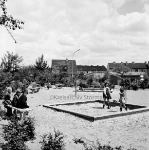 Kellerberg-Süd: Kinderspielplatz in Neubaugebiet: links drei Kinder auf Bank: rechts zwei Kinder in Sandkasten: hinten Klettergerüst, weiterer Sandkasten und Bänke: dahinter Büsche: hinten zwei Wohnblocks und ein Einfamilienhaus