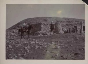 Hugo Grothe zu Pferd : Expeditionsleiter und Geograph Hugo Grothe (1869-1954) auf dem Pferd. Neben ihm ein Mann, im Hintergrund Bewohner vor einem Haus.