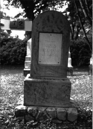 Grabstein 72 (Index: 100101526) Material: Sandstein, weiße Marmorplatte, Rundbogen mit Kupferumkleidung verstärkt Zustand: Stele gesprungen Maße HxBxT: 129x39x21 cm Beschreibung: Stele, getreppter Sockel, unterer Sockelteil rusticaartig, Rundbogenabschluß, rechteckige Schriftplatte: Grabstein 72