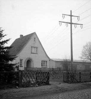Überlandleitung: Starkstrommast neben Wohnhaus: vorn Tanne und Gartenzaun: hinten rechts Wirtschaftsgebäude