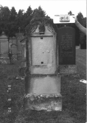 Grabstein 24 (Index: 99609660) Material: Sandstein Zustand: verwittert, Schrifttafel fehlt Maße HxBxT: 156x53x27 cm Beschreibung: Stele, Abschluß Sonderform, rechteckige eingelassene Schrifttafel fehlt, getreppter Sockel Symbolik: Vase mit zwei Zweigen: Grabstein 24