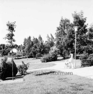 Grünanlage zwischen Markt, Matthias-Claudius-Schule und Ahrensböker Straße: Rasen, Büsche, Bäume, Frau mit Kinderwagen auf Fußweg: rechts Terrasse mit Jägerzaun, Straßenlampe: links Matthias-Claudius-Gedenkstein
