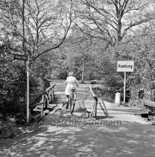 Lottbek: An der Lottbek: provisorisch ausgebesserte Holzbrücke über die Lottbek Richtung Hamburg: darauf Mädchen mit Fahrrad: links Straßenschild: rechts Ortsschild von Hamburg: dahinter Büsche und Bäume: hinten rechts Pkw
