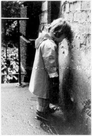 Kleines Kind mit Regenjacke steht an eine Mauer gelehnt (Altersgruppe 18-21)