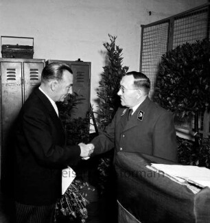 Reichsbund, Kreisverband: Ehrung: Kreisvorsitzender Gustav Bomann mit Blumenstrauß in Hand bei Händedruck mit Postbeamtem: vorne Rednerpult: hinten links Metallspinde, rechts Metallgitter, Kübelpflanzen