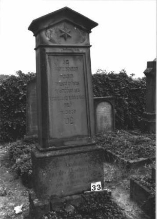 Grabstein 33 (Index: 99808473) Material: Sandstein Zustand: verwittert Maße HxBxT: 200x65x28 cm Beschreibung: konische Stele, getreppter Sockel, unterster Sockelteil rusticaartig, giebelartiger Abschluß mit Verdachung, rechteckiges Schriftfeld eingetieft Symbolik: seckszackiger Stern: Grabstein 33