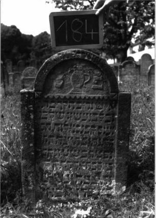 Grabstein 184 (Index: 100103364) Material: Sandstein Zustand: stark verwittert Maße HxBxT: 80x55x12 cm Beschreibung: Stele, eingezogener Rundbogenabschluss, Schriftfeld rechteckig eingetieft, oben durch Zackenfries begrenzt, im Bogen Levitenkanne Symbolik: Levitenkanne, Zackenfries: Grabstein 184