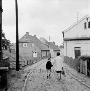 Kirchsteig: Kopfsteinpflaster: Strommast: niedrige Häuser, Gartenzäune: in Straßenmitte Frau mit Junge an der Hand