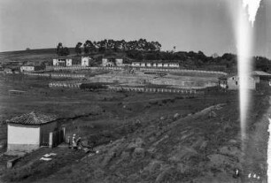 Fazenda : Blick auf eine Fazenda mit Wirtschaftsgebäuden und einer Kapelle im Mittelgrund. Im Vordergrund vier Personen neben einem kleinen Gebäude an einem Bach. Links auf dem Feld ein Fußballtor.