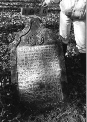 Grabstein 1026 (Index: 99751285) Material: Sandstein Zustand: stark verwittert Maße HxBxT: 120x66x8 cm Beschreibung: Stele, geschwungener Abschluß, Schriftfeld eingetieft Symbolik: Medaillon: Grabstein 1026