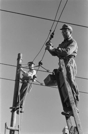 Energieversorgung. Arbeiten an einer Oberleitung. Ansicht mit zwei Arbeitern (auf Mast und von einer Leiter aus arbeitend)