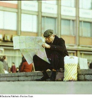 Alter Mann mit Mütze sitzt auf einer niedriger Mauer und liest im Stadtplan