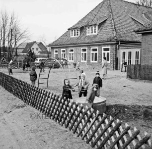 Kindergarten in ehemaligem Volksschulgebäude: Schulstraße: vorn Hof mit Spielgerät und spielenden Kindern: rechts hinten Aufsichtsperson: hinten links Pkw, Bäume und Häuser: im Vordergrund Jägerzaun
