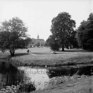 Schlosspark: äußerer Schlossgraben mit Seerosen, Schwan: Spaziergänger auf Fußweg, Frau auf Parkbank, Bäume: im Hintergrund Gottesbuden, Schlosskirche