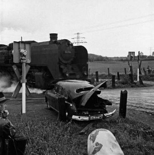 Siebenbäumen (Kreis Herzogtum Lauenburg): Bahnstrecke zwischen Bad Oldesloe und Ratzeburg: Bahnübergang der Landstraße zwischen Bad Oldesloe und Ratzeburg: Filmaufnahmen der Reginald Puhl Filmproduktion aus Hamburg-Wandsbek zum Dokumentarfilm "Todesfallen": nachgestellter Bahnunfall einer Dampflokomotive mit einem Pkw: vorn Köpfe von Zuschauern: in der Mitte beschädigter Pkw: dahinter Bahnübergang mit Dampflokomotive: links und rechts Signalanlagen: hinten rechts Wiese