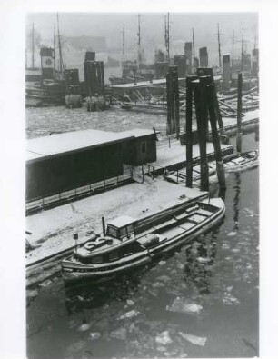 Hamburg. Der Hafen im Winter. Eine Barkasse, zum Transport von Hafen- bzw. Werftarbeitern gedacht, liegt im teilweise mit Eisschollen bedeckten Hafenbecken. Im Hintergrund liegt die Flottille der Schlepper bereit, ihre Arbeit zu verrichten