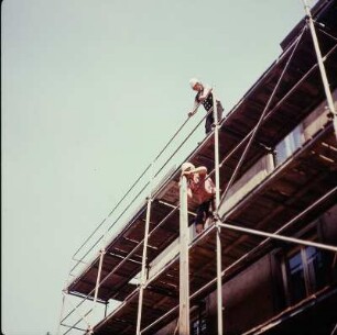 Zimmerer auf einem Gerüst beim vertikalen Weiterreichen einer Holzbohle