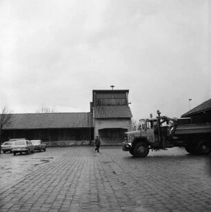 A 1: Bad Oldesloe / Rethwischfeld: Autobahnmeisterei: hinten Betonsilo für Streugut, an Fassade Wanduhr: daneben Lagerhallen: auf Hof parkende Pkws, rechts automatischer Streu-Lkw: Mann beim Überqueren von Hof