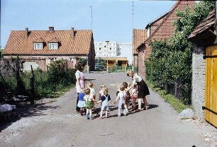 Berlin. Zwei Kindergärtnerinnen mit einer Gruppe von Kindern auf einem Spaziergang zwischen Siedlungshäusern. Blick auf Neubau-Wohnblöcke im Hintergrund