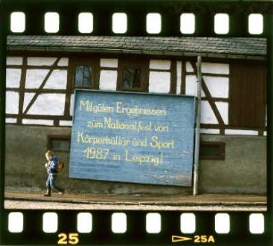 Zum Nationalfest von Körperkultur und Sport : Transparent vor einem Haus an dem ein kleiner Junge vorbei geht