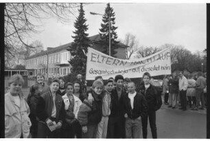 Emil-Nolde-Schule [Grundschule]: Am Schulzentrum: Elternprotest gegen Umwandlung in Gesamtschule: Schülergruppe, hinten Eltern mit Transparent "Eltern wacht auf. Gesamtschule ja - auf diese Art nein": hinten Schulgebäude, Flachbau: 20. März 1990