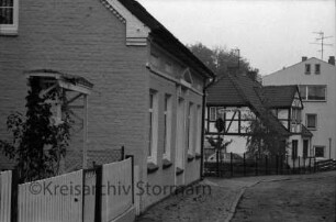 Straße mit Kopfsteinpflaster: in der Mitte Gehweg: links und hinten Zäune und Wohnhäuser