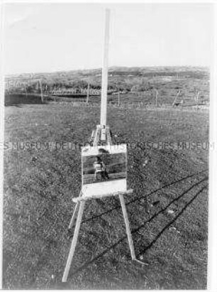 Staffelei in der Landschaft mit dem Bild einer sitzenden Person vor dieser Landschaft (Altersgruppe 18-21)