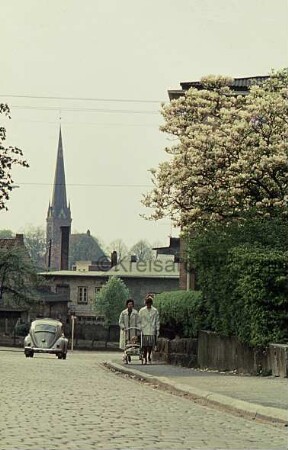 Ratzeburger Straße mit Kopfsteinpflaster: links Pkw: rechts zwei Frauen mit Kinderwagen vor Mauer mit Hecke und blühendem Magnolienbaum: hinten Mitte Einmündung der Bahnhofstraße: dahinter Gebäude: hinten links Turm der Peter-Paul Kirche