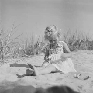Kleines Mädchen mit Kleid sitzt und spielt am Strand