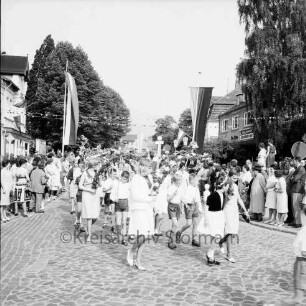Kindervogelschießen: Umzug: Mädchen mit Blumenbögen, Jungen mit Bänderstäben, beim Abbiegen in andere Straße: Fahnenschmuck, Wimpelgirlande über Straße: Zuschauer an Straßenrändern: Bäume, 26. Juni 1965