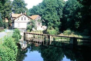 Wehr in einem Flusslauf; im Hintergrund Großbäume und Gebäude
