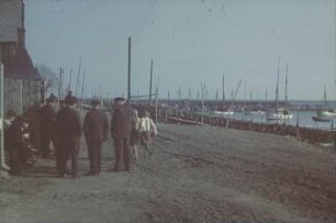 Reisefotos. Kleiner Hafen mit Fischerbooten. Am Ufer zum Trocknen ausgebreitete Fischernetze und Gruppe von Männern (vielleicht in Holland)