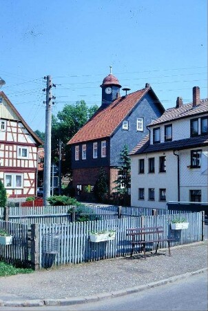 Teich am Dorfplatz mit Staketenzaun, dahinter alte Schule