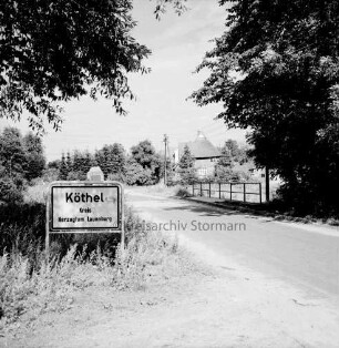 Köthel (Kreis Herzogtum Lauenburg): Ortseingang mit Ortsschild: im Hintergrund rechts Bauernhaus, links Ehrenmal: hinten Bäume