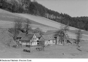 Ein Gehöft am Fuße eines Hügels (vermutlich in Thüringen), im Hintergrund ein Wald