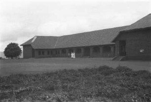 Schule von Peramiho : Blick auf ein großes Gebäude in einer Landschaft mit drei Personen davor.