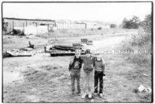 Drei Jungen in einer Reihe, die Arme umeinander gelegt, dahinter Freifläche mit Sperrmüll (Prämiertes Foto Sonderthema: Spiele spielen)